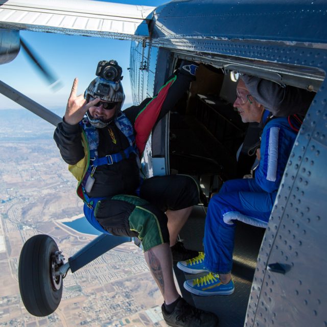 tandem skydiving videographer hangs on outside of airplane and poses for the camera looking excited for the skydive